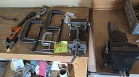 Assorted clamps and vices displayed on wooden workbench. Several C-clamps, a spring clamp, a drill press vice unattached, and a large bench vice bolted to the bench.