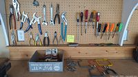 Pegboard with assorted hand tools including locking pliers, adjustable wrenches, standard pliers, and screwdrivers arranged on hooks above a wooden surface with a box containing small nails and screws, plus loose C-clamps and hardware.
