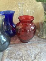 Close-up of several vases showing a cobalt blue glass vase, medium red vase, and other clear and colored glass vases in various shapes.