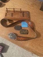 Photo of vintage brown leather belt with three metal belt buckles including Coors branded buckles and wooden valet tray on wooden surface.