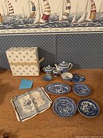 Blue Willow doll dinnerware set displayed with box and cloth napkin on a wooden surface against nautical themed wallpaper.