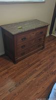 Front and side view of a brown wood dresser with three drawers and dark metal handles. A removable stone top with natural marbled pattern is fitted on top. Positioned against wall on wood floor.