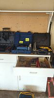 Wide view showing the cabinet with black DeWalt case holding the power drill, blue Drill Doctor 400 case, drawer opened showing assorted drill bits, and a blue empty DeWalt case on the countertop.