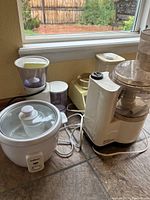 Wide view of four electric kitchen appliances on table by window showing Salton rice cooker, Braun grinder, Regal blender, and food processor with cords.
