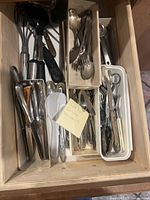 Drawer open showing multiple trays of flatware and kitchen utensils including spoons, forks, knives, ladles, and peelers.