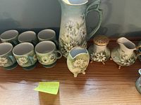Floral ceramic dishes including coffee cups, pitchers, creamer, napkin holder, and shaker shown on wooden surface.