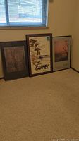 Three framed landscape posters leaning against wall beneath a window on carpeted floor