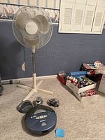 Roomba vacuum and Holmes pedestal fan positioned on carpeted floor, surrounding area with boxes and wall decor