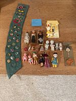Top-down view of dolls and Girl Scout sash on wooden floor, showing assortment of dolls and badges on sash.