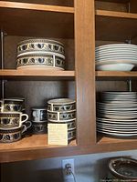Photo of stacked plates, bowls, and mugs with the Mikasa Intaglio pattern in wood kitchen cabinet.