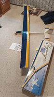 Model airplane laid flat on carpeted floor showing top wing surface with blue and yellow color scheme, and part of the fuselage and tail visible. Box and manual nearby.