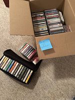 Box containing a large quantity of music CDs, organized vertically with covers visible. Adjacent to the box are cassette tapes, some in a black zippered case and others loose.