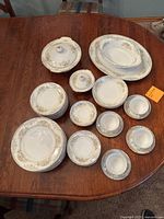 Photo shows the set of dishes including various plates, sugar bowl with lid, covered casserole dish, platter, and serving dish arranged on a table. Floral pattern with gold trim visible.