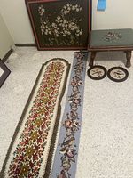 Photo showing small needlepoint ottoman, floral framed picture, two needlepoint runners, narrow bird wall hanging, and two small round bird pictures arranged on floor and wall