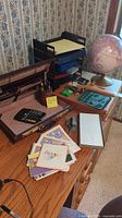 Full desk view showing briefcase open with interior compartments, calculator, pen holder, greeting cards, and a stack of desk organizers with yellow notepad plus a world globe on stand.