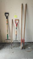 Four garden tools leaning against the wall: 5 tine manure fork, 4 tine digging fork, garden tiller with long handles, and pitch fork