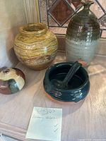 Photo showing three pottery items: large beige and brown layered vase, small two-toned round vase, and dark green soup crock with lid and ladle.