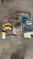 Full view of assorted hand saws, power saws, jigsaw, and blades on concrete floor.