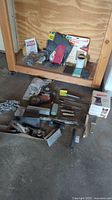 Overview of the garage items showing assorted metal files, sandpaper, sanding blocks, a woodwork finisher hand tool, and other woodworking accessories laid out on a wooden surface and concrete floor.