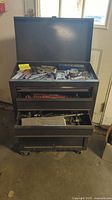 Gray metal rolling tool chest with top lid open showing assorted metal tools and parts inside.