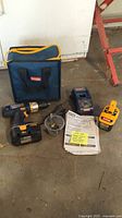 Full kit arranged including drill, batteries, charger, drill bits in clear container, instruction manuals, and carrying case placed on a concrete floor with a white wall background.