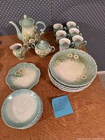 View of the tea set showing teapot with lid, creamer, sugar bowl, six cups, six saucers, six plates, and an additional flower plate. Creamer has visible glue repair.