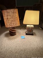 Full view of two small lamps side by side: left lamp with spiral wood base and textured shade; right lamp with porcelain floral base and pleated shade, both plugged in and illuminated.