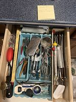Drawer of kitchen utensils showing various graters, scoops, corkscrews, and other food preparation tools in a blue plastic tray.
