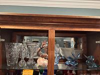 Wide view of collection inside wooden hutch showing various crystal and art glass vases, bowls, and figurines including blue art glass duck and compote, dolphin vase, and a pink flower figurine.