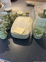 Photo showing 6 rectangular plates stacked, mugs, creamer, sugar bowl, and salt and pepper shakers with green winter landscape design on a dark countertop.
