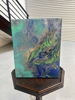 Front view of abstract acrylic painting in green and blue tones resting on a dark wooden surface.