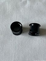 Pair of black ear plugs and tunnels shown side by side on white fabric background. Items have glossy black finish and hollow center.