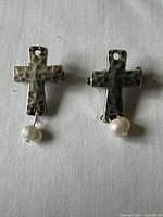 Front view of two silver-tone cross brooches each with a single hanging white pearl-like bead on white fabric background.