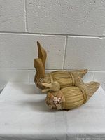 Two vintage handmade wood and reed duck decoys placed on a white surface against a white brick wall. One duck has a carved wooden head raised upward, and the other has a ribbon with a small rose tied around its neck.