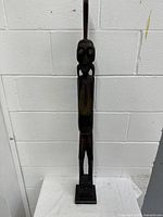 Full frontal view of the tall wooden statuette showing entire figure and base against white brick wall.