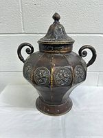 Front view of the antique vintage urn showing the detailed embossed medallions, scroll handles, lid with finial, and attached chain.