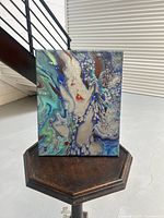 Front view of abstract canvas painting on a small wooden stand, showing the swirling blue, teal, white, gray, and red patterns.
