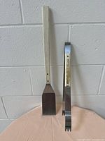 Two BBQ accessories standing against a white cinderblock wall on a beige surface: a metal spatula with a wooden handle and curved metal tongs with a wooden handle, both approximately 21 inches long.