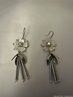 Pair of flower earrings with translucent petals and iridescent center stones, showing dangling silver-tone metal tubes with leaf-shaped charms on a neutral background.