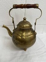 Front view of round brass teapot with wooden handle and removable lid, showing wear and tarnish on surface.