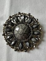 Front full view of the round brooch showing central faceted cabochon, teardrop dark rhinestones, and decorative antique silver-tone metal filigree.