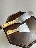 Both scraper spatulas laid flat on a wooden surface showing full shape and size difference.