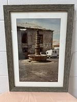 Front view of decorative wooden photo frame with rustic textured grayish wood and white matte border showcasing a photo of a stone fountain and building on a street.