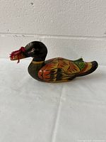 Front and side view of the wooden duck decoy showing bright colorful painted feathers and red tassel in beak.