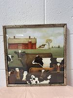 Framed picture showing a farm scene with multiple cows and a red barn in the background, hung on a white cinder block wall.