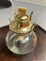 Top angled view of the glass and brass oil lamp showing the clear textured glass body and brass wick holder with flat wick and adjustment knob.