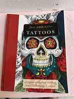 Front cover of hardcover coloring book showing tattoo skull with floral designs and text indicating over 120 illustrations.