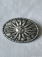 Front of oval pewter lapel pin showing sun face with radiating rays and star cutouts.