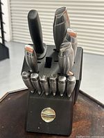 Front view of KitchenAid wooden knife block holding multiple knives with metal and black plastic handles, showing some wear and one broken blade