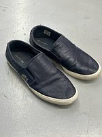 Pair of black Lacoste leather slip-on shoes on a gray floor showing top and outer side. Shoes show some creasing and signs of use. White rubber soles are visible.